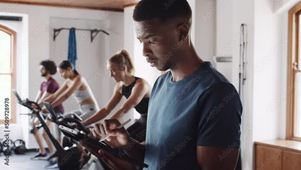 Young adult black male typing on electronic tablet at an indoor fitness gym. Confident fitness professional providing guidance. Young adult black male typing on electronic tablet at an indoor fitness gym. Confident fitness professional providing guidance.