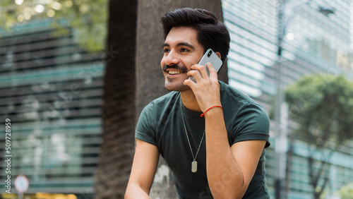 Latin man with mustache smiling, sitting on a bench making a phone call.