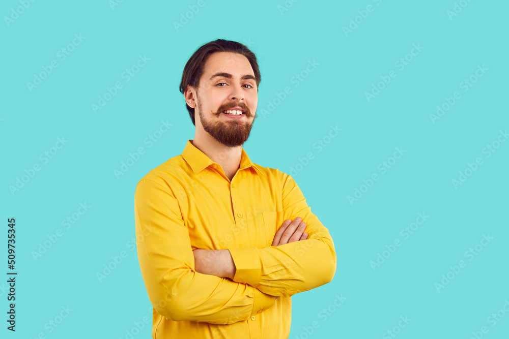 Portrait of handsome and happy man with stylish mustache and beard on light blue background. Confident millennial Caucasian man in yellow shirt stands with folded hands and smiling looking at camera. Portrait of handsome and happy man with stylish mustache and beard on light blue background. Confident millennial Caucasian man in yellow shirt stands with folded hands and smiling looking at camera.
