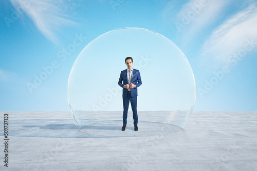 Attractive young european businessman standing under and inside big glass sphere cover on concrete ground and bright blue sky background. Protection and limitation concept.