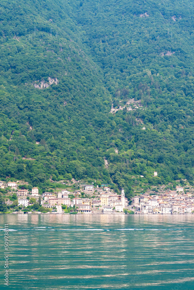 Vertical view of a typical ancient italian village on the coastline of Lake Como, Lombardy, Italy. Turquoise rippled waters in the foreground. Green trees on the background.