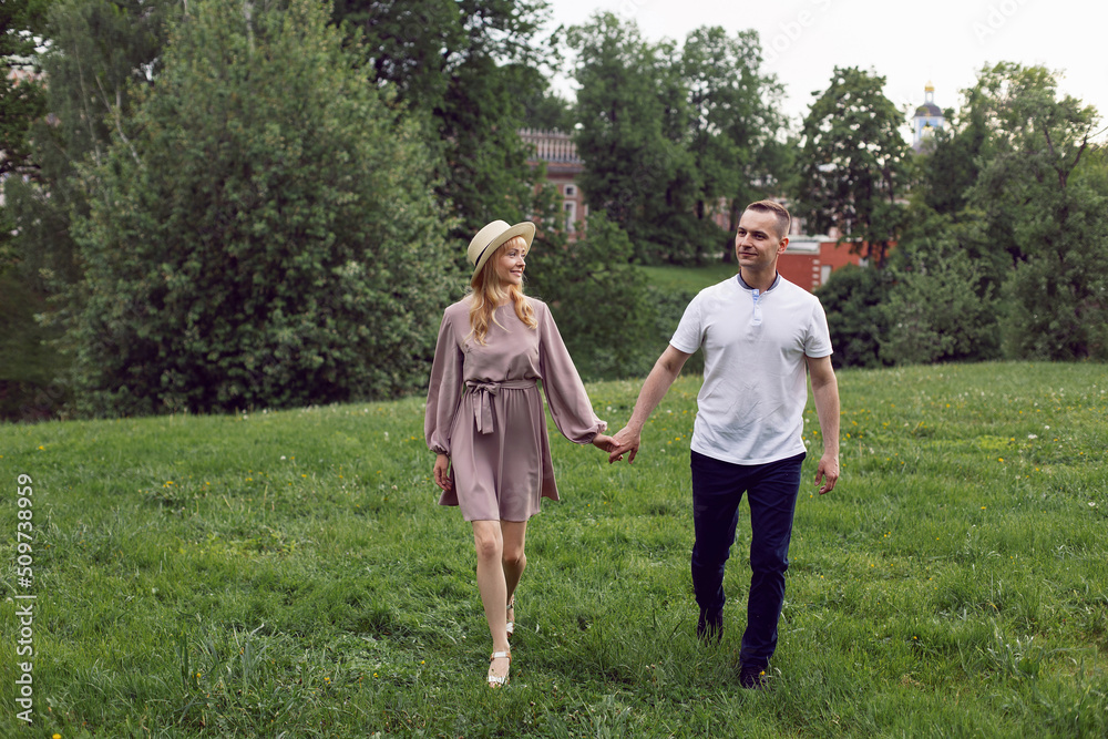 man and a woman in love in a dress and hat are walk on a green field under a tree in summer.