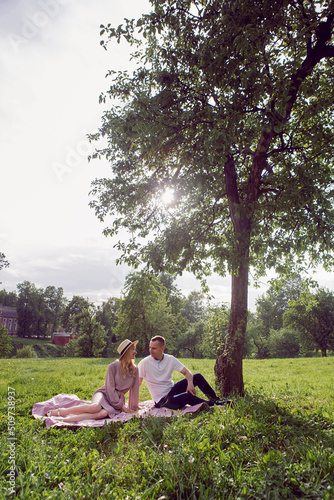 man and a woman in love in a dress and hat are sitting on a blanket on a green field under a tree in summer. © saulich84