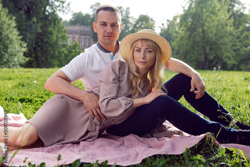 man and a woman in love in a dress and hat are sitting on a blanket on a green field under a tree in summer.