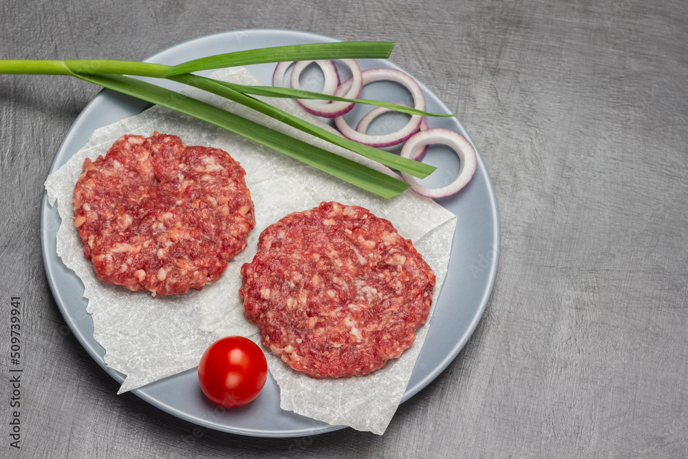 Two raw beef burgers with green onion and tomato on gray plate. Two raw beef burgers with green onion and tomato on gray plate.