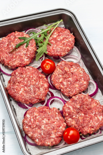 Raw beef burgers with chopped onions and tomatoes on baking tray.