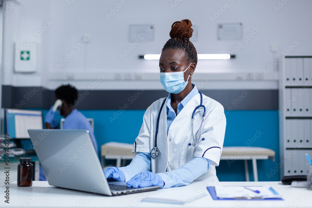 Polyclinic general practitioner expert using laptop to check patient record files. Polyclinic specialist prescribing medication while wearing virus protection facemask because of covid pandemic. Polyclinic general practitioner expert using laptop to check patient record files. Polyclinic specialist prescribing medication while wearing virus protection facemask because of covid pandemic.