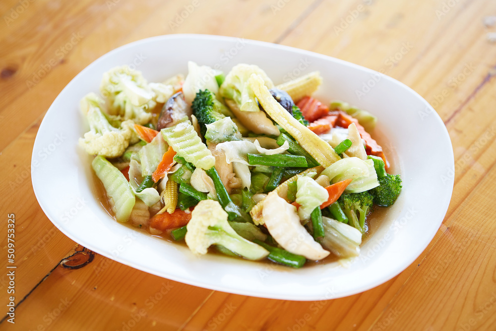 Stir Fried Mixed Vegetables (Sliced baby corn, oringi mushrooms, shiitake, carrots, kale, cauliflower, cauliflower and yard long beans) are served with white plate on wooden table. good healthy food Stir Fried Mixed Vegetables (Sliced baby corn, oringi mushrooms, shiitake, carrots, kale, cauliflower, cauliflower and yard long beans) are served with white plate on wooden table. good healthy food