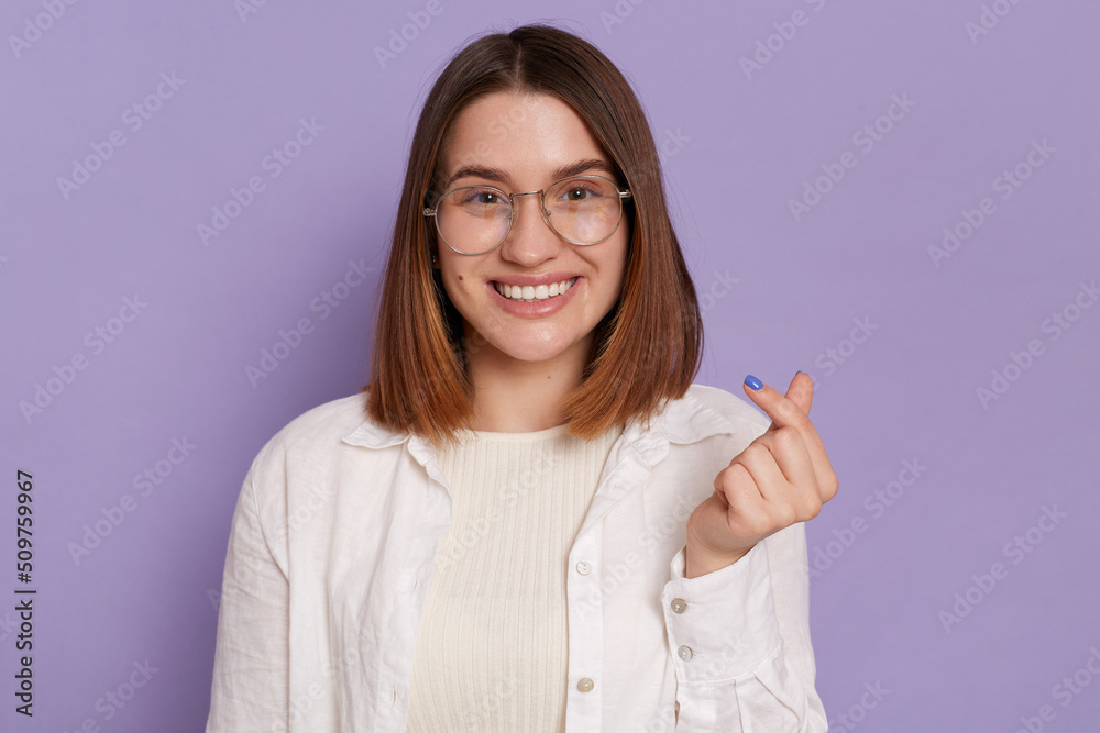 Pretty dark haired woman with gentle smile makes korean like sign expressing love to you, wearing white shirt, posing isolated over purple background. Miniheart gesture. Pretty dark haired woman with gentle smile makes korean like sign expressing love to you, wearing white shirt, posing isolated over purple background. Miniheart gesture.