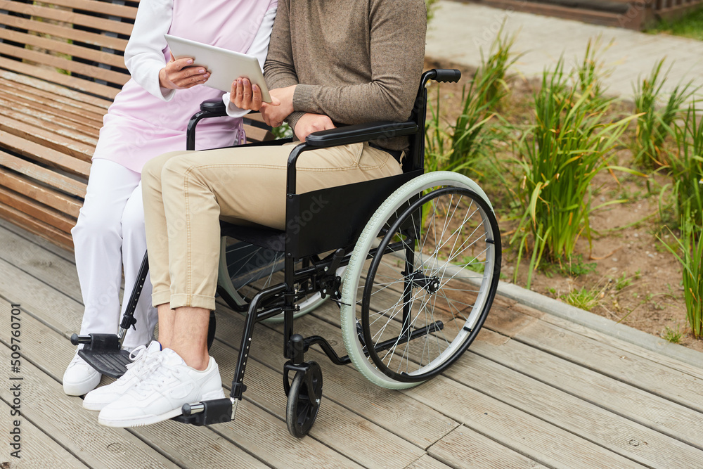 Obraz premium Unrecognizable nurse in uniform sitting on bench in yard of hospital and showing video on tablet to handicapped man in wheelchair