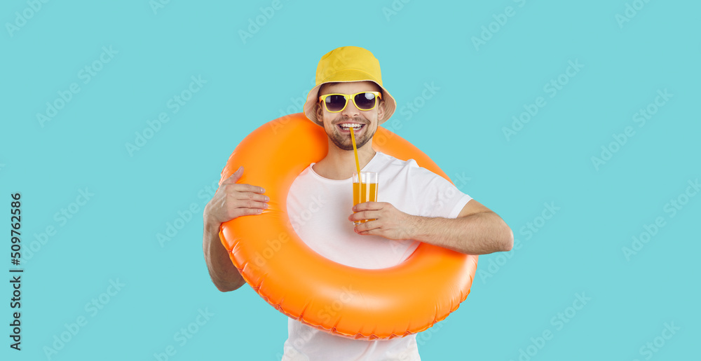 Happy young man with inflatable swimming circle and with glass of juice enjoys summer vacation. Cheerful funny guy in summer clothes drinks juice on light blue background. Summer relaxation concept. - obrazy, fototapety, plakaty 