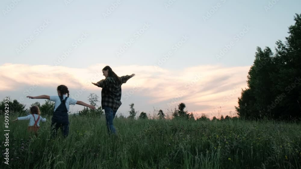 Happy family enjoying park at sunset. Teamwork, freedom, summer weekend concept. Loving fun young mother and smiling playful two little daughters kid are run green grass on background colorful sky Happy family enjoying park at sunset. Teamwork, freedom, summer weekend concept. Loving fun young mother and smiling playful two little daughters kid are run green grass on background colorful sky