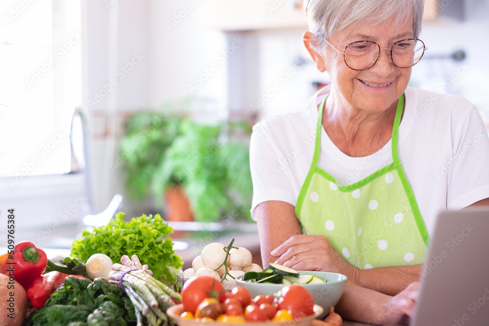 Attractive senior woman preparing vegetables in the home kitchen browsing on laptop for new recipes. Caucasian elderly people enjoying healthy vegetarian eating