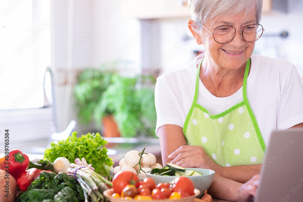 Attractive senior woman preparing vegetables in the home kitchen browsing on laptop for new recipes. Caucasian elderly people enjoying healthy vegetarian eating Attractive senior woman preparing vegetables in the home kitchen browsing on laptop for new recipes. Caucasian elderly people enjoying healthy vegetarian eating