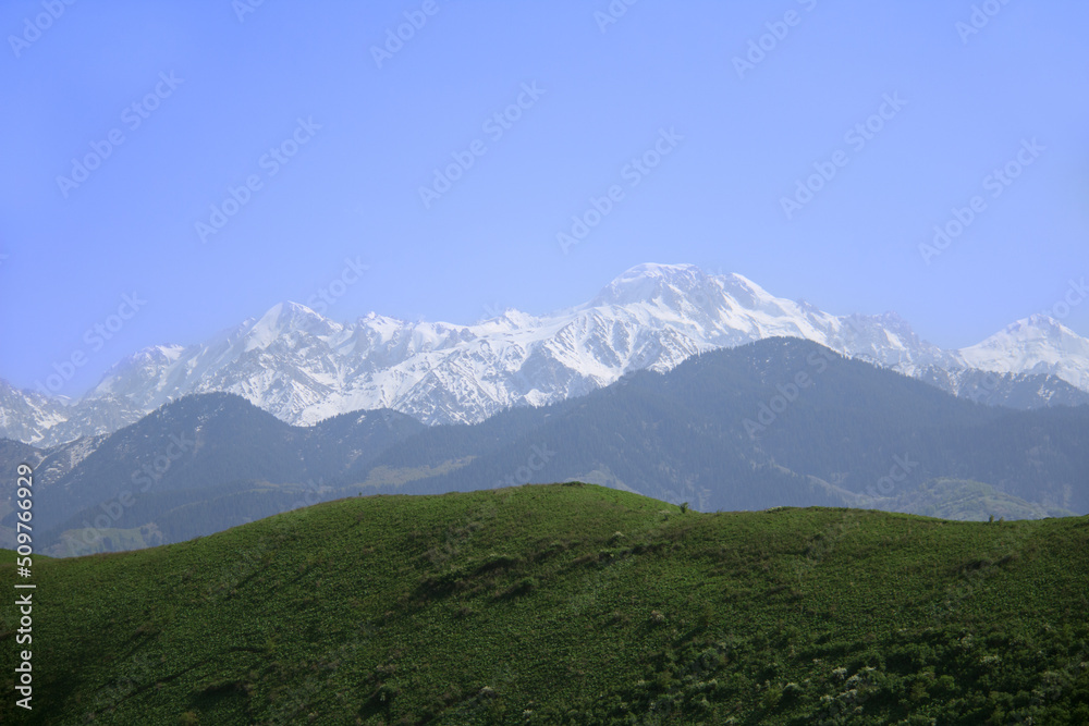Snow-covered alpine ridge, Talgar peak in the middle, another ridge below and large green hills, clear sky, spring, sunny