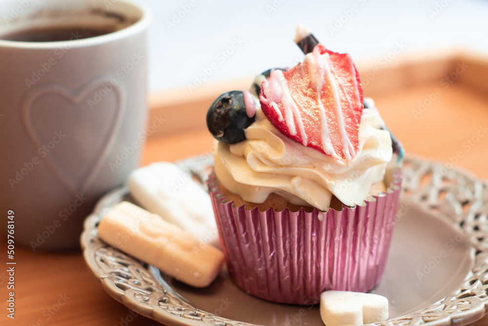 Delicious dessert and coffee in a heart dessin mug on a tray. Creamy muffin cupcake decorated with strawberry and sweets - desaturated, moody colors.