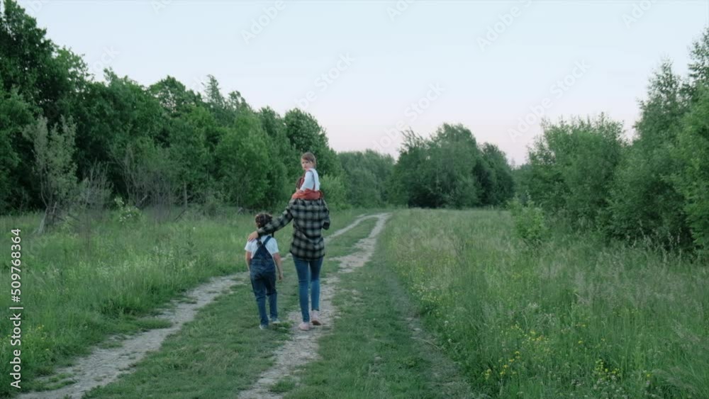 Selective focus of loving young mother with two daughter child walking on road on background sky and forest. Slow-motion. Caring mom and cute little girls kids enjoying nature together, outdoors Selective focus of loving young mother with two daughter child walking on road on background sky and forest. Slow-motion. Caring mom and cute little girls kids enjoying nature together, outdoors