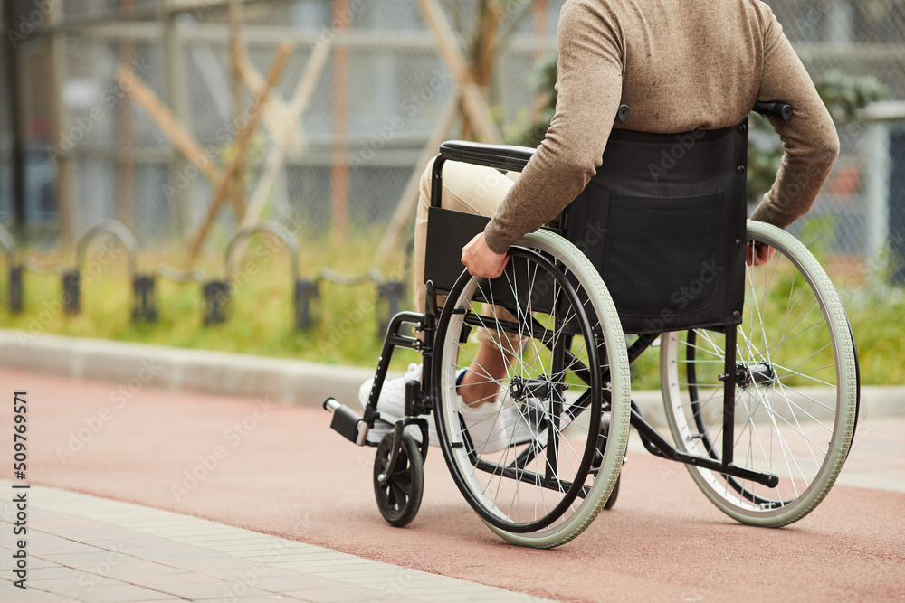 Unrecognizable disabled man in brown sweater sitting in modern wheelchair and turning wheels of wheelchair with hands while walking alone Unrecognizable disabled man in brown sweater sitting in modern wheelchair and turning wheels of wheelchair with hands while walking alone