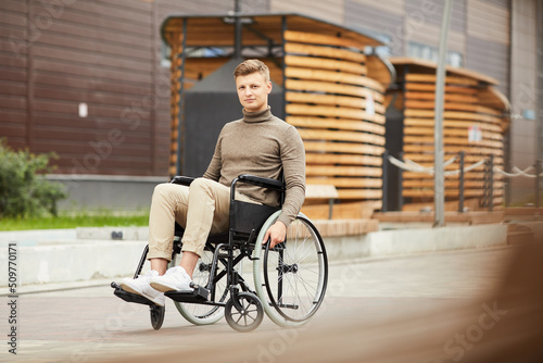 Content handsome young guy with spinal cord injury sitting in wheelchair outdoors and looking at camera