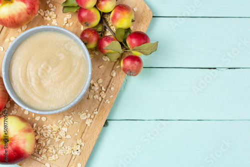 Oatmeal porridge for the baby from ground cereals in a blue bowl, red ripe apples on a wooden board on a blue background. Space for text. Baby nutrition, the first complementary feeding of a child.