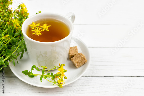 A drink (infusion, decoction) of St. John's wort in a cup on a saucer and a piece of brown sugar on a white wooden background. Herb and flowers of the medicinal plant Hypericum. Space for text.