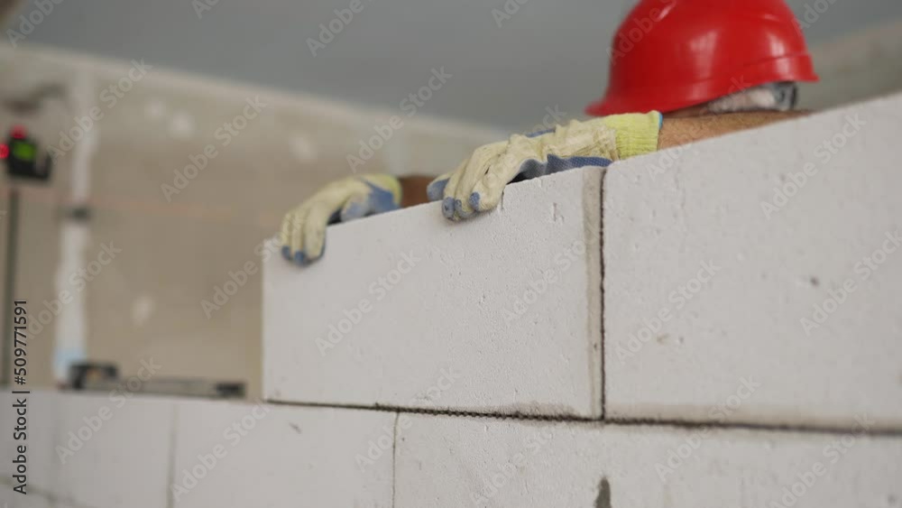 Bricklayer or mason lays bricks to construct wall of aerated concrete blocks. Brickwork worker contractor taps foam concrete bricks with mallet for precise level masonry on construction site.