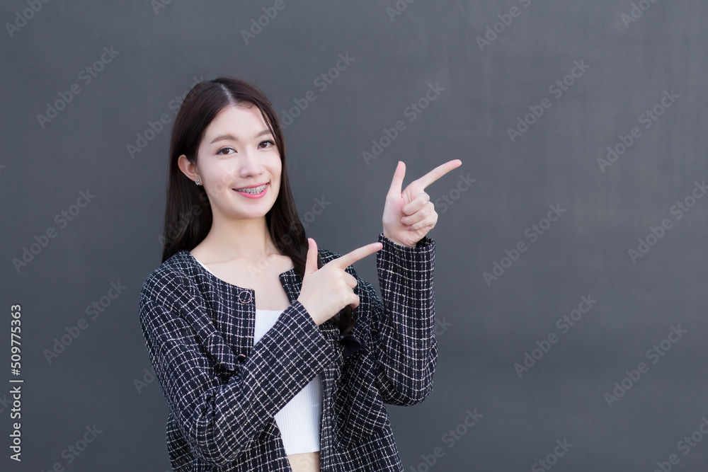 Asian working woman who wears black suit with braces on teeth is pointing hand to present something on the dark gray wall. Asian working woman who wears black suit with braces on teeth is pointing hand to present something on the dark gray wall.
