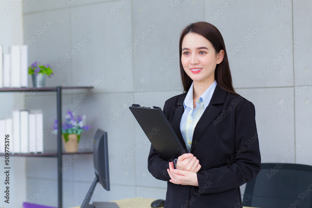 Professional Asian business young woman in black suit smiles happily while she works and holds clipboard in office. Professional Asian business young woman in black suit smiles happily while she works and holds clipboard in office.