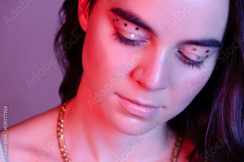 Portrait of young dreamy woman in neon lights with closed eyes and gold shiny creative stars make-up on her eyes.