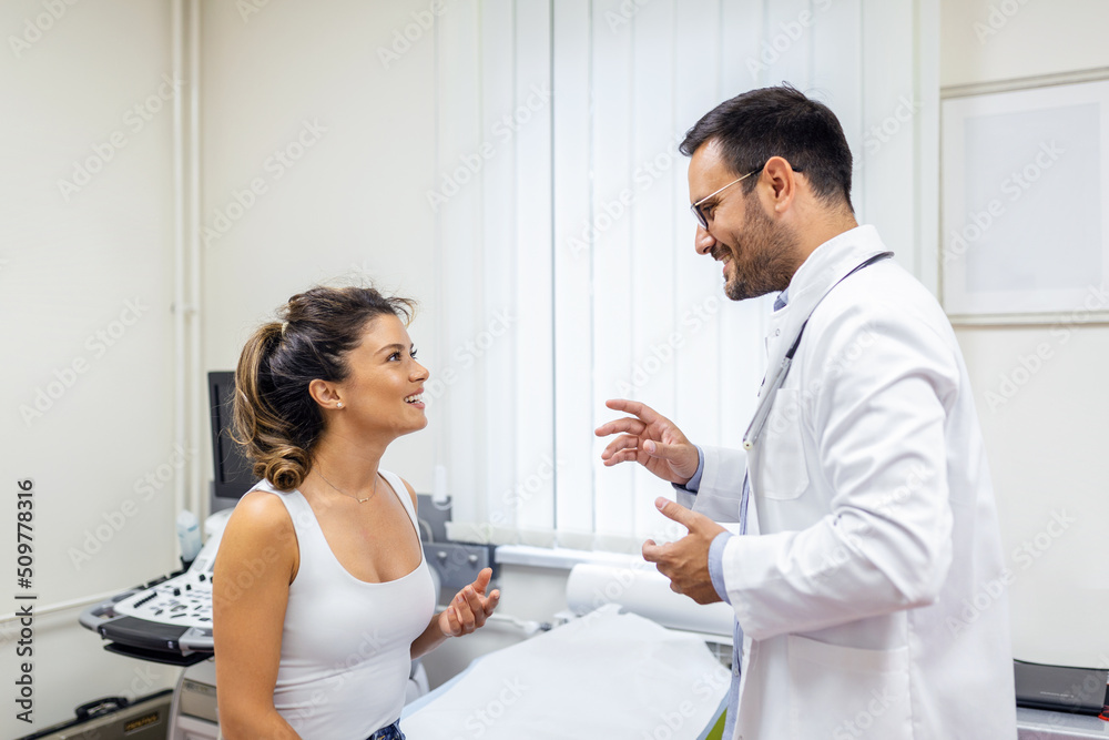 Fototapeta premium Male doctor consulting female patient at consultation. Professional physician wearing white coat talking to woman at appointment visit in clinic.