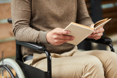 Close-up of unrecognizable man in casual outfit sitting in wheelchair outdoors and reading book about mental health of disabled people © Mediaphotos