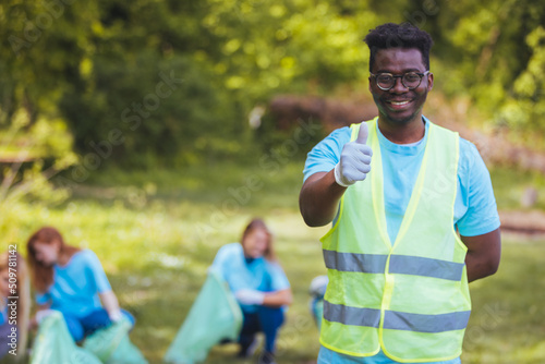 A young African man smiles at the camera while volunteering with his neighbors to pick up garbage in their neighborhood park. He is holding a garbage bag. Thumb up...