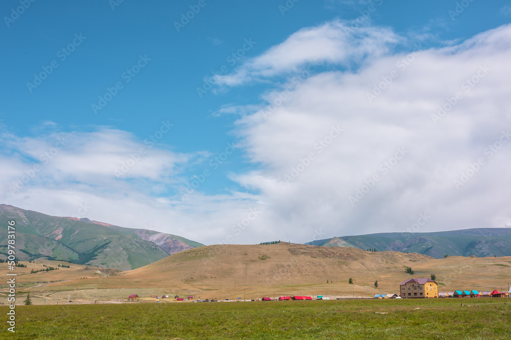 Dramatic alpine landscape with small houses in sunlit green meadow against large hill under clouds in blue sky. Beautiful mountain scenery at changeable weather. Scenic view to high mountain range.