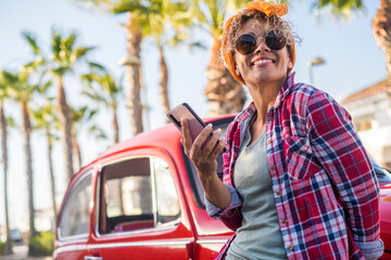 Cheerful trendy young adult woman use app mobile phone to plan and set next travel destination traveling with a red classic car in background. Happy people and transport vehicle concept leisure