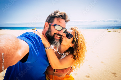 Adult couple of tourist take selfie picture at the beach with sand and blue ocean and sky in background. Hapy man and woman enjoying summer holiday vacation together. Travel lifestyle people photo