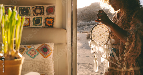 Boho woman traveler standing on the door of her alternative home camper van holding a dreamcatcer. Daydreaming lifestyle concept with free female people with scenic background place