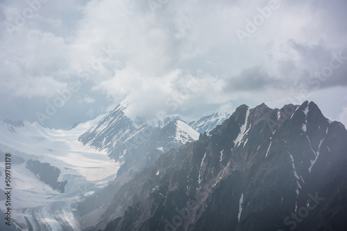 Atmospheric landscape with fuzzy silhouettes of sharp rocks and snowy mountain top in low clouds during rain. Dramatic view to large snow mountains and glacier blurred in rain haze in gray low clouds.