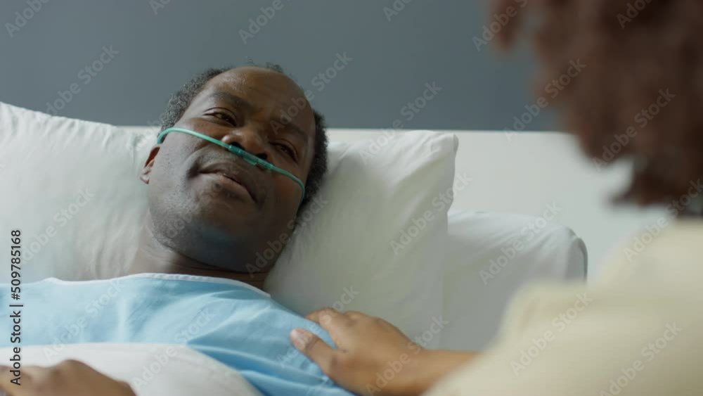 Tilt up over the shoulder shot of sick African American man with nasal cannula lying on hospital bed and talking with loving wife as she holding his hands and stroking shoulder Tilt up over the shoulder shot of sick African American man with nasal cannula lying on hospital bed and talking with loving wife as she holding his hands and stroking shoulder