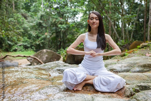 Asian young female lady yoga model meditate and practicing yoga zen poses in a nice, quiet tranquil green outdoor forest park with giant stone, tree and nice little stream waterfall