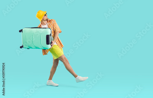 Funny young man who is ready for summer vacation having fun walking with suitcase on light blue background. Joyful guy in summer clothes holding suitcase near copy space. Full length. Web banner. © Studio Romantic