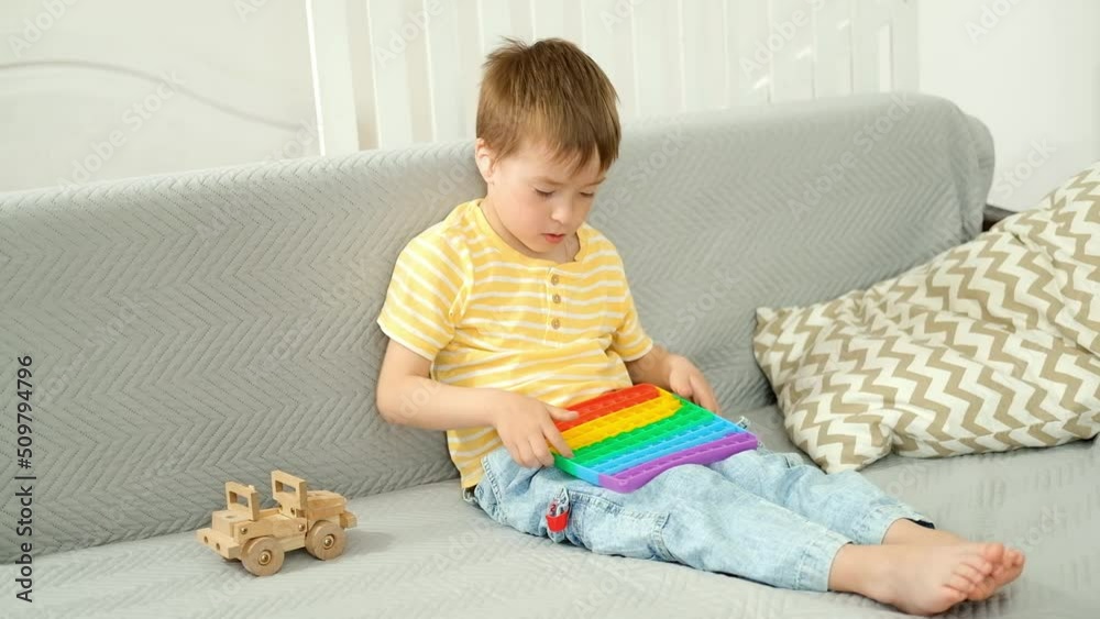 Caucasian boy is sitting at home in living room on sofa and playing with an antistress pop it toy. Caucasian boy is sitting at home in living room on sofa and playing with an antistress pop it toy.