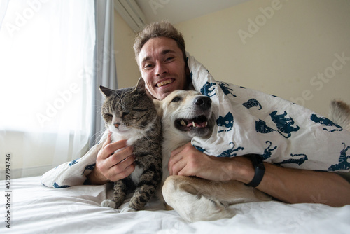 Pleased young man lying in bed with dogs and cat