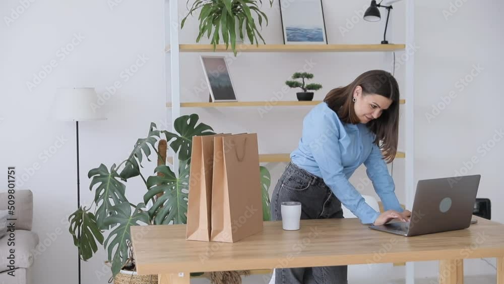 Happy businesswoman working in a home office standing looking at her laptop computer surrounded by gift bags and packaging for the products for sale on her new website