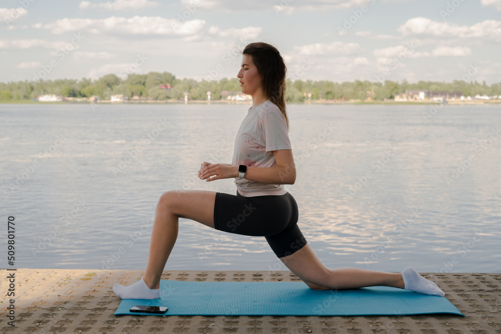 Fototapeta premium a young girl is doing yoga on the shore on the yoga mat of the river in the evening