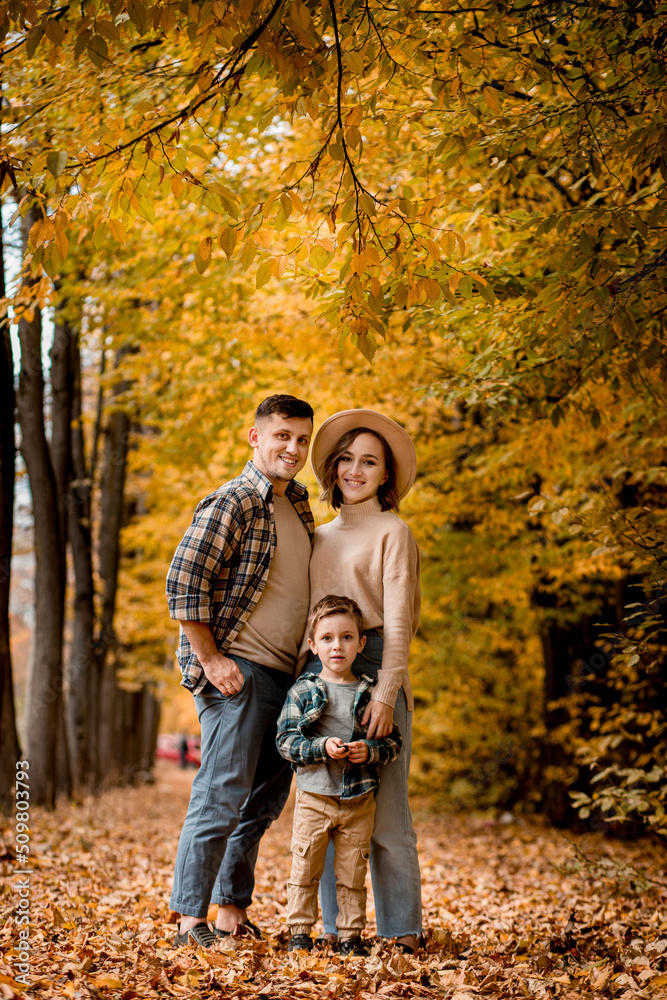 Fototapeta premium Portrait of happy young family on the background of autumn park. Mom, dad and son are smiling. Family autumn photo shoot