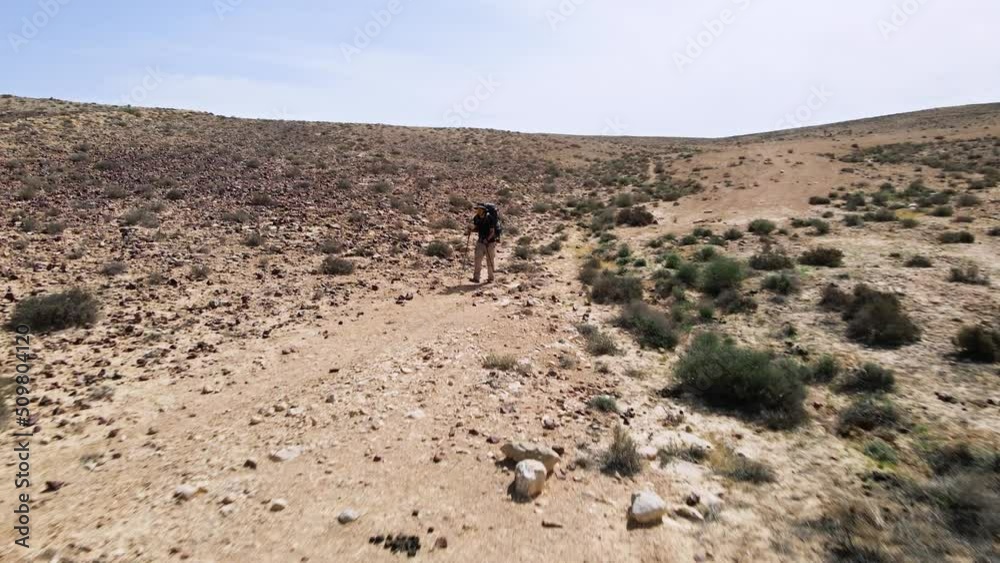 Fast drone fly in towards hiker walking a track in rocky desert wilderness landscape. Hiker has full equipment with poles and backpack. Location is Ramon crater in the Negev Desert, Israel Fast drone fly in towards hiker walking a track in rocky desert wilderness landscape. Hiker has full equipment with poles and backpack. Location is Ramon crater in the Negev Desert, Israel
