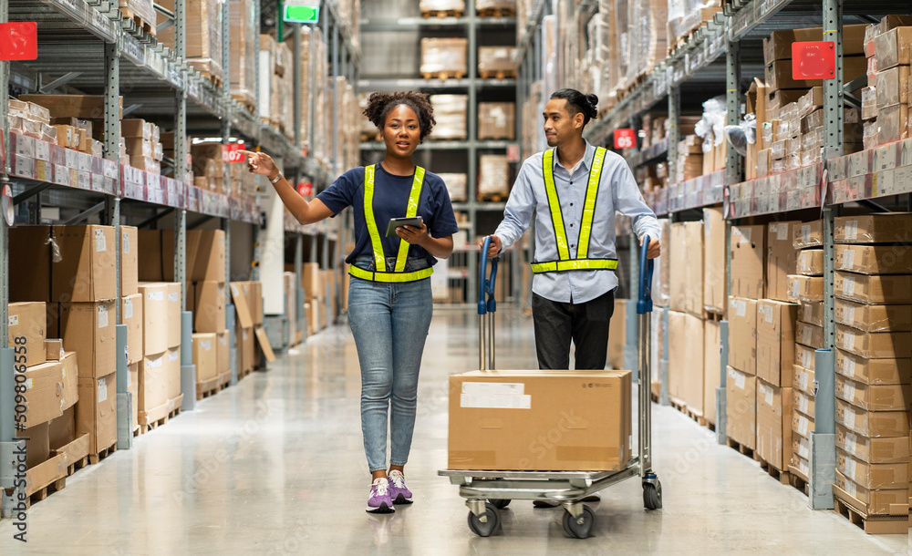 African American woman walks with a tablet and an Asian man picks up boxes in a cart to deliver to customers walking in a warehouse at a furniture wholesaler.