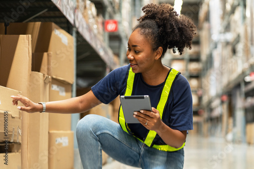 African-American woman walks with a tablet counting furniture items on a shelf in a furniture wholesale warehouse.