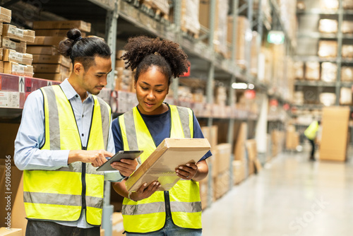Asian male worker carries a tablet to count furniture items and an African American woman carries a box inspecting bracode before sending it to customers in a furniture wholesale warehouse.