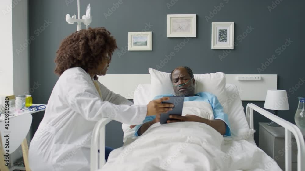 Zoom in shot of African American female doctor sitting at bedside in hospital ward and discussing something on digital tablet with male patient